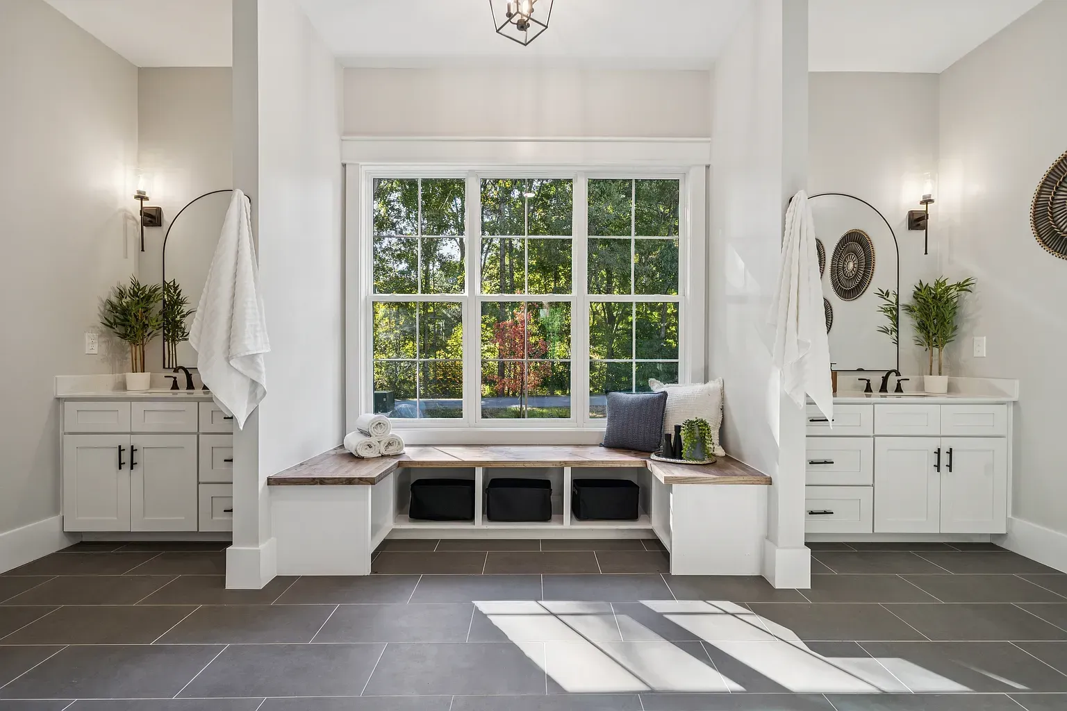 Bright bathroom with window seat, twin vanities, gray tile floor, and sunlight streaming in.