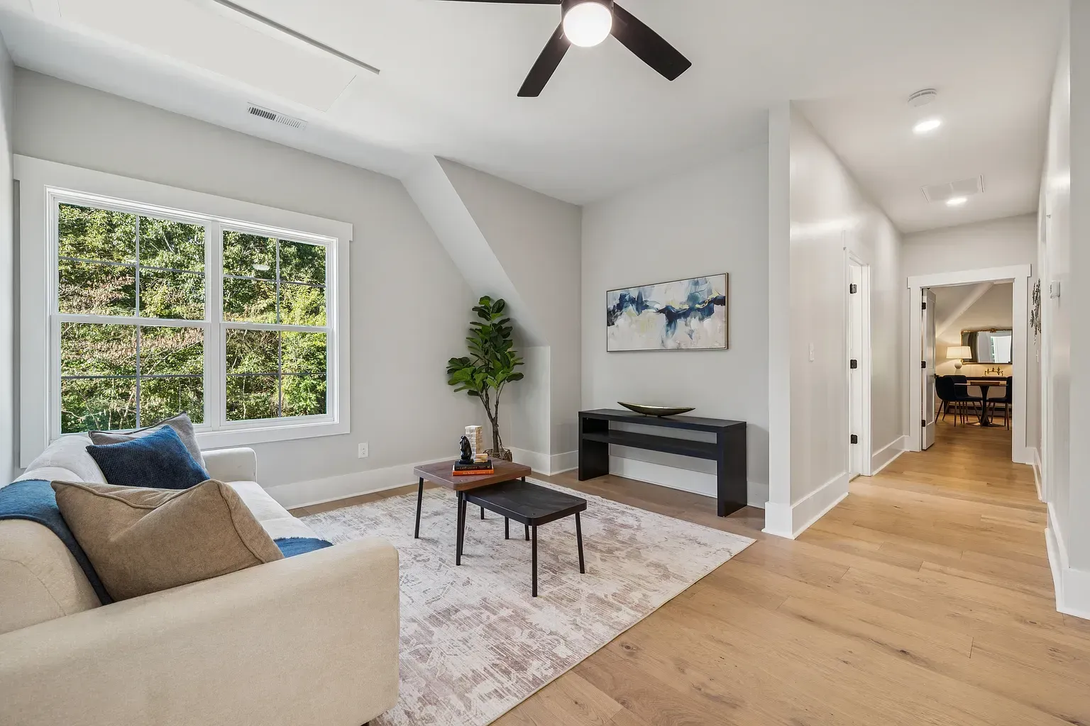 Bright modern living room with beige sofa, blue pillows, coffee table, TV console, and large window with greenery.