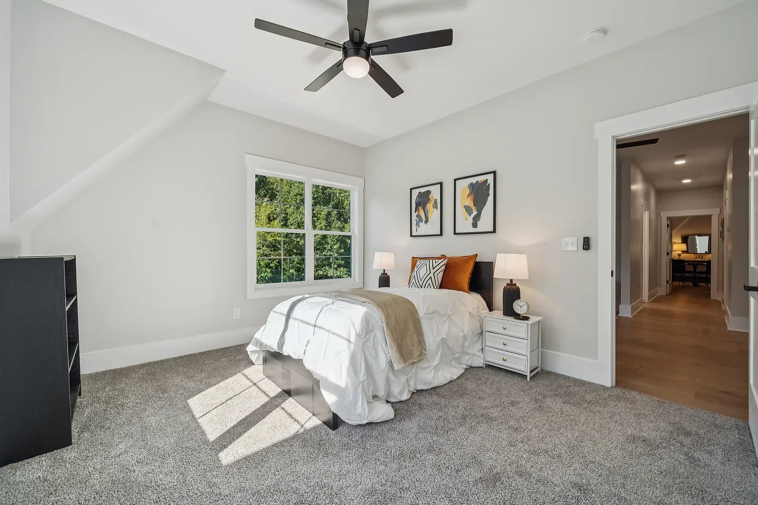 Bright bedroom with white bedding, gray carpet, ceiling fan, and an open doorway to a hall.