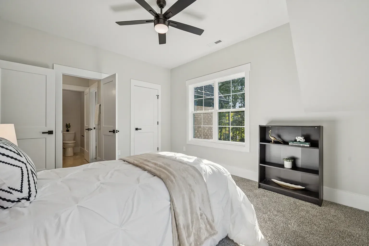 Bright bedroom with white walls, bed, ceiling fan, window, and dark shelving unit near the wall