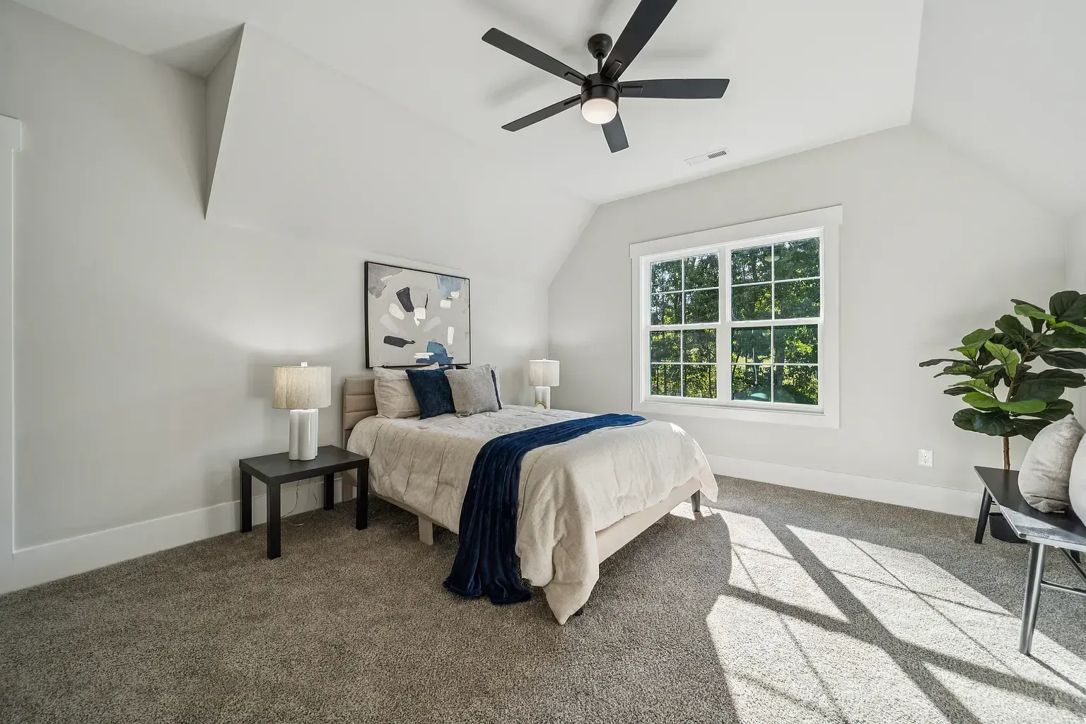 Bright bedroom with gray carpet, white walls, bed, ceiling fan, and sunlit window.