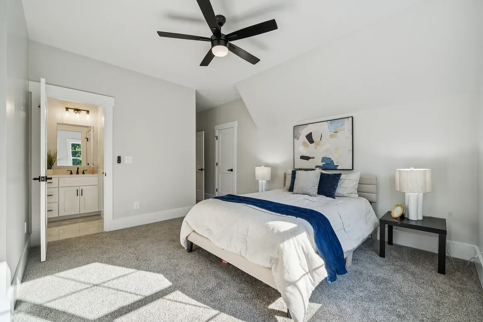 Bright bedroom with a bed, nightstands, ceiling fan, and an open doorway to a bathroom.