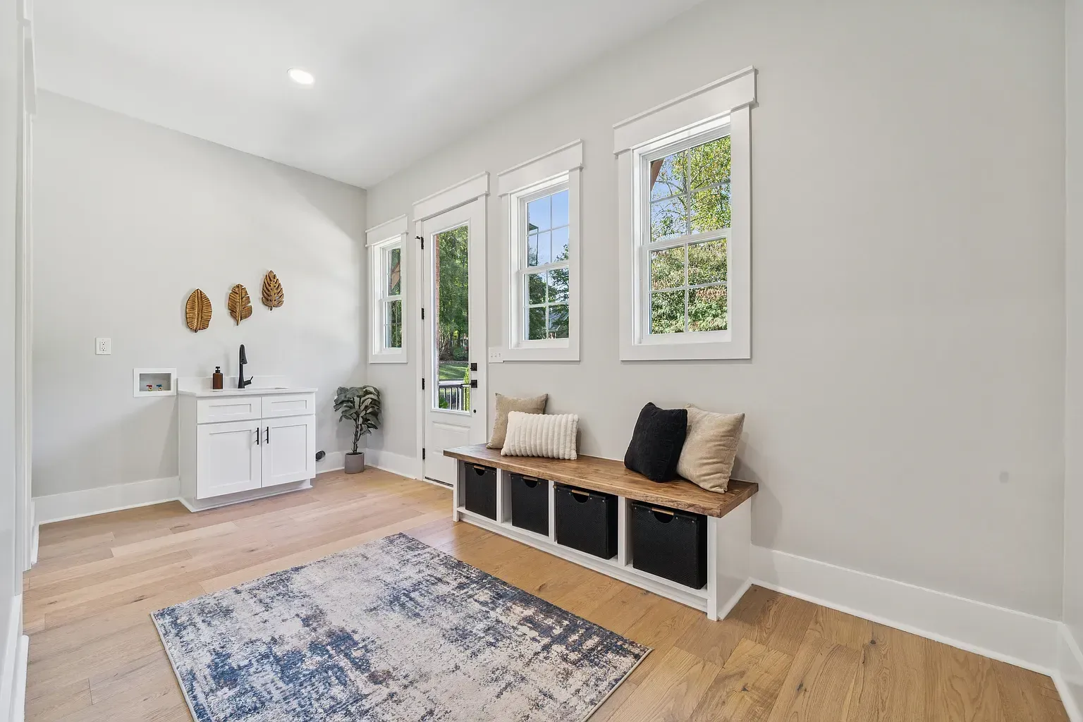 Bright white living room with wood floors, windows, rug, bench, and small cabinet.