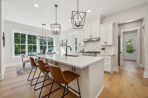 Bright white kitchen with island seating, gold fixtures, and hardwood floors, viewed from the dining area.
