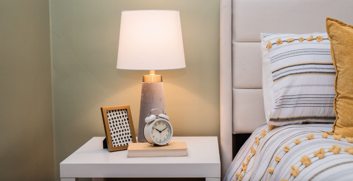 Bedroom nightstand with lamp, alarm clock, and framed photo beside a neatly made bed with patterned pillows