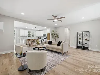 Bright modern living room with white sofa, armchairs, rug, shelving, and open kitchen in the background