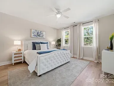 Bright beige bedroom with a white bed, nightstand, large rug, and windows with curtains