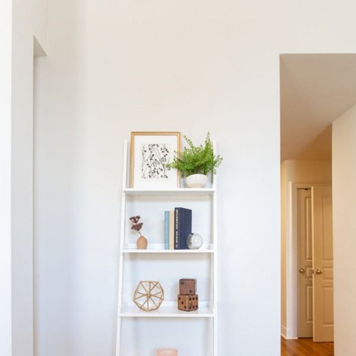 White wall with a small shelf holding books, framed art, a plant, and decor beside a hallway and door.