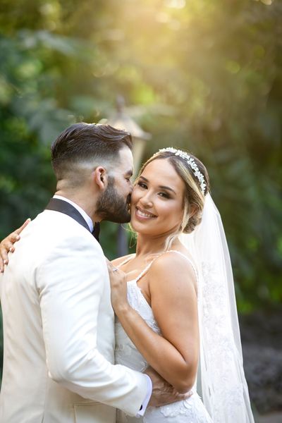 Bride and groom embracing outdoors, smiling under soft sunlight with a veil and white wedding dress