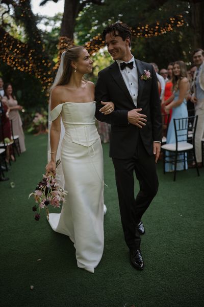 Bride and groom walking arm-in-arm outdoors at a wedding reception, smiling under trees