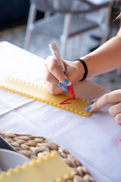 Hands drawing with a red pencil on white fabric at a table, with blue yarn and a yellow ruler nearby