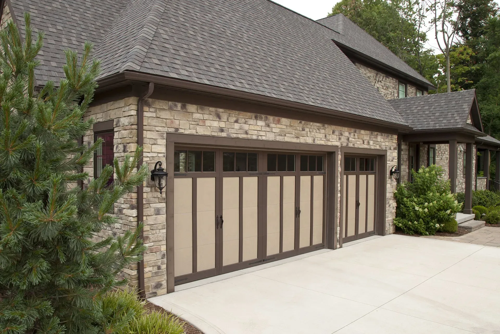 Stone-faced house with tan garage doors, brown trim. Concrete driveway, green bushes, and trees surround the structure.