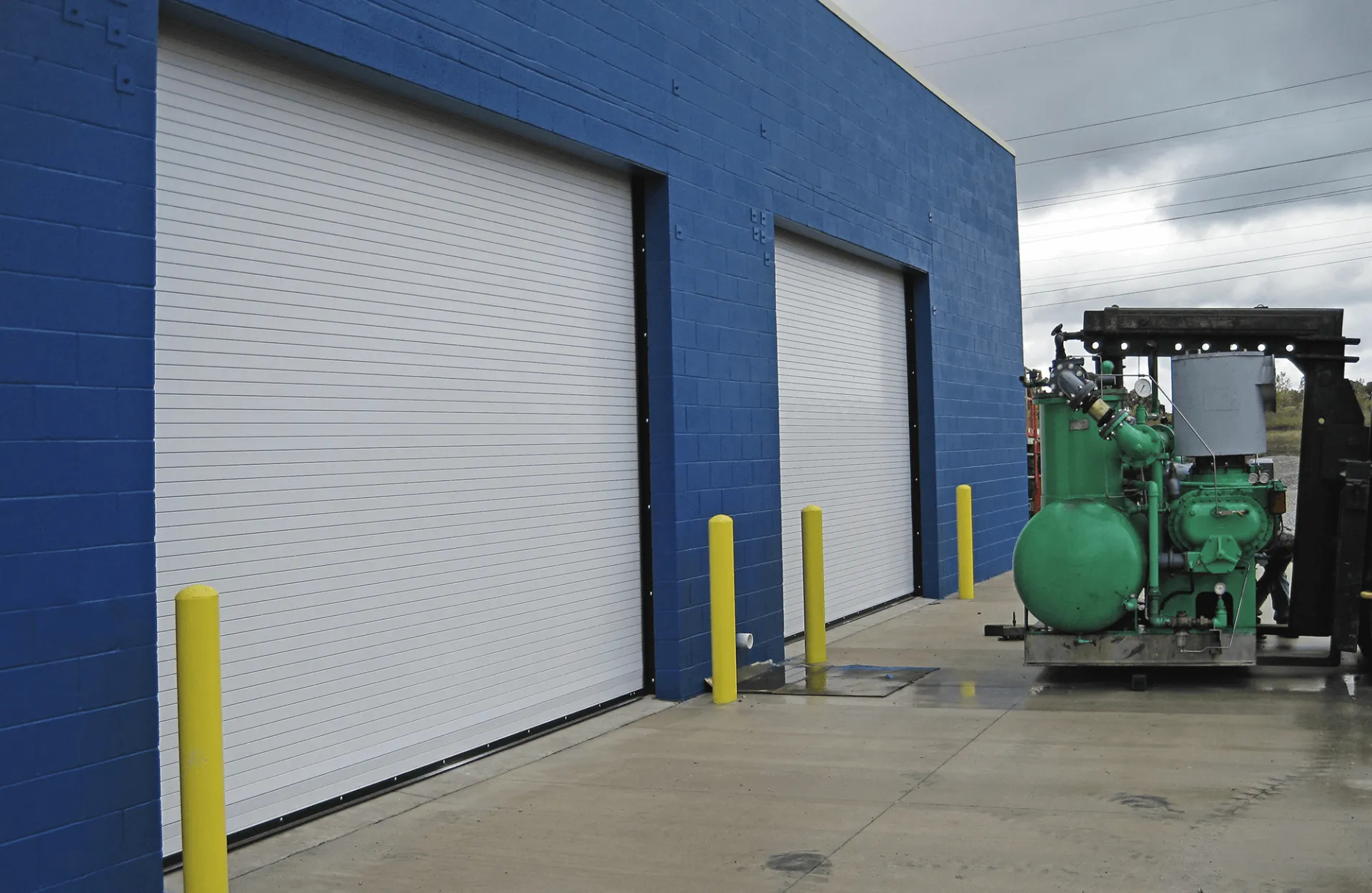 .Large Garage Door Commercial Blue building with two white roll-up doors, yellow bollards, and machinery outside.