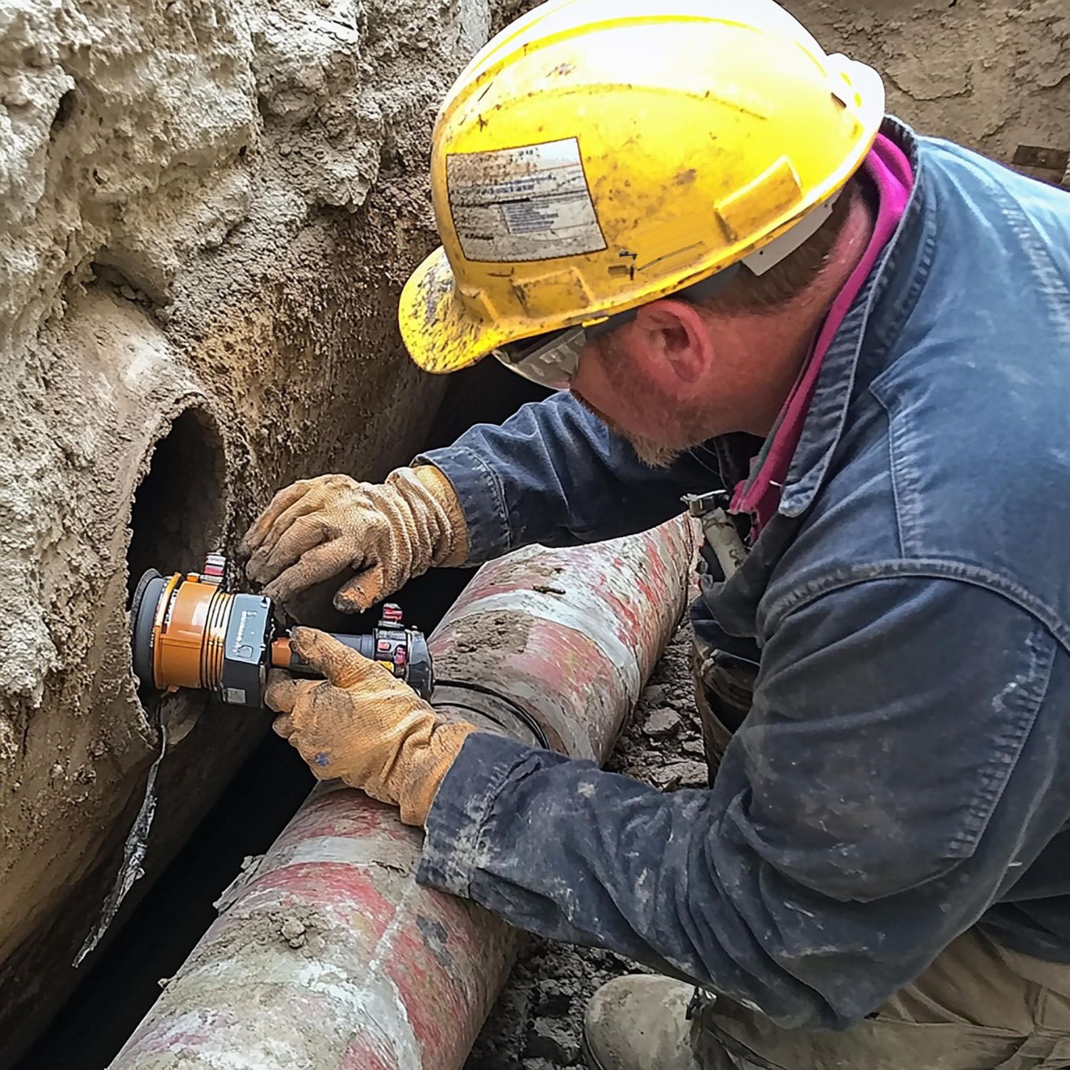 A worker in a yellow hard hat and gloves inspects an open pipe in a dirt trench.