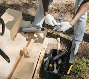 Person wearing gloves handles wood beside a chainsaw and toolbox outdoors in a work area
