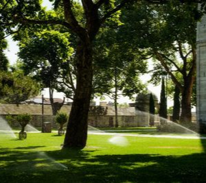 Lawn with sprinkler spray under trees beside a stone building on a sunny day