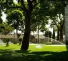 Lawn with sprinkler spray under trees beside a stone building on a sunny day