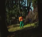 Person in orange shirt and green pants walking in a wooded area behind a fence.