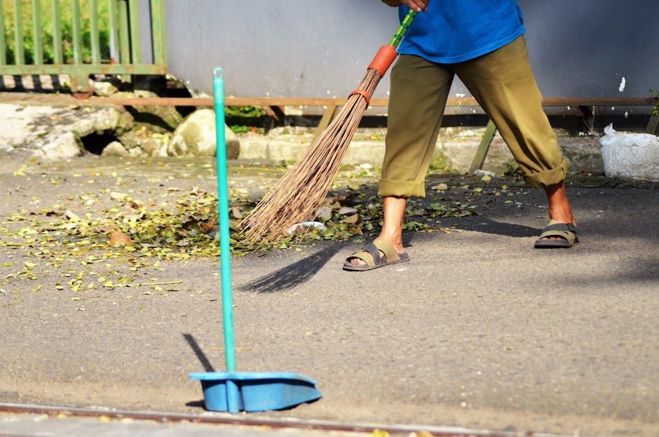Worker sweeping pavement with a broom next to a blue dustpan on a street