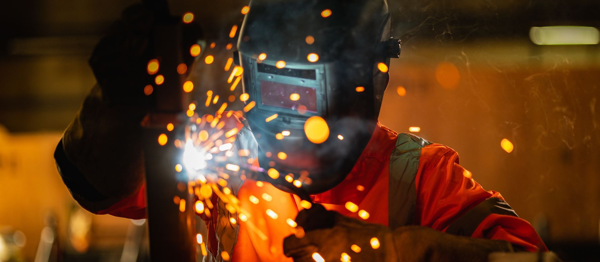 A worker welding metal with sparks flying, safety gear glowing in dim industrial light.]