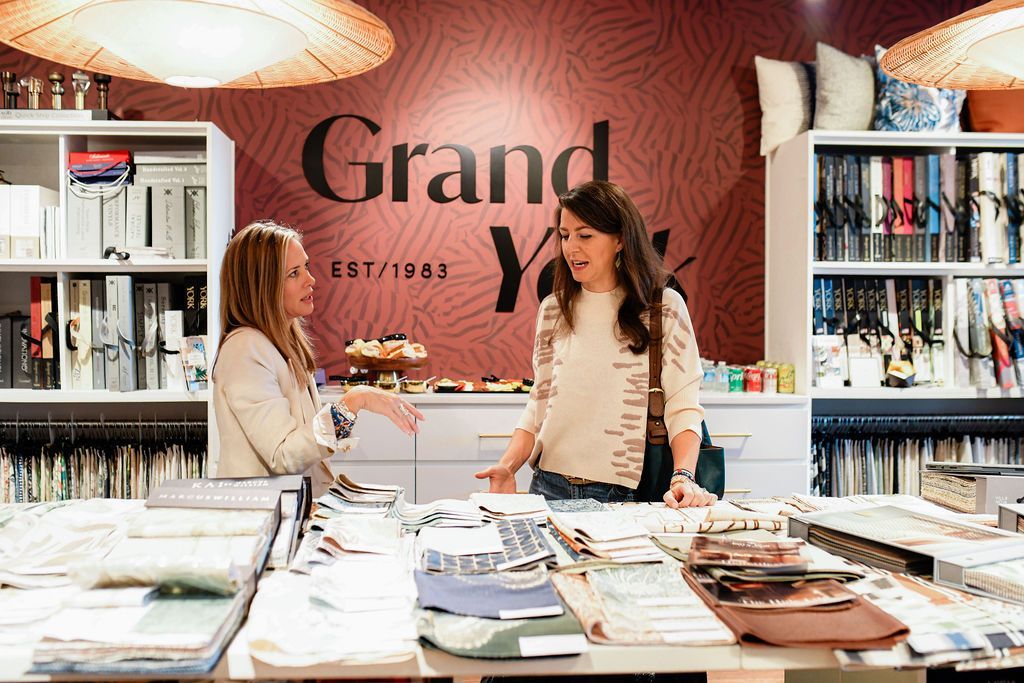 Two women at a design shop counter, looking at fabric samples. The store sign says