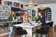 Three people at a design store counter, examining fabric samples. Shelves of textiles and lamps are in the background.