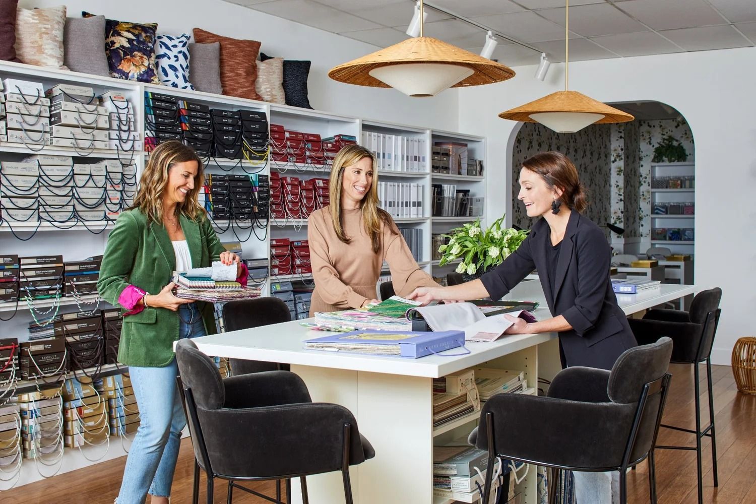 Three people at a design store counter, examining fabric samples. Shelves of textiles and lamps are in the background.