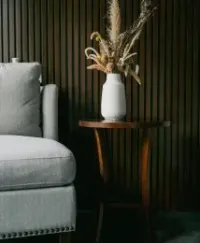 Gray armchair next to a small wooden table with a white vase holding dried flowers against a brown paneled wall.