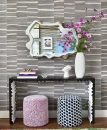 A console table with a mirror and vase, flanked by patterned stools against a striped gray wall.