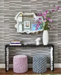 A console table with a mirror and vase, flanked by patterned stools against a striped gray wall.