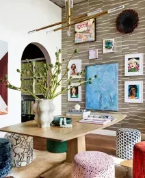 Room with a wooden table, colorful stools, and art-filled wall. Includes a green plant and an archway with books.