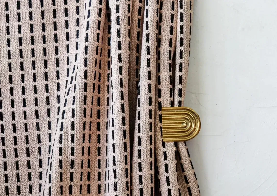 Beige curtain with black stitch pattern held by a gold, half-circle curtain tieback.