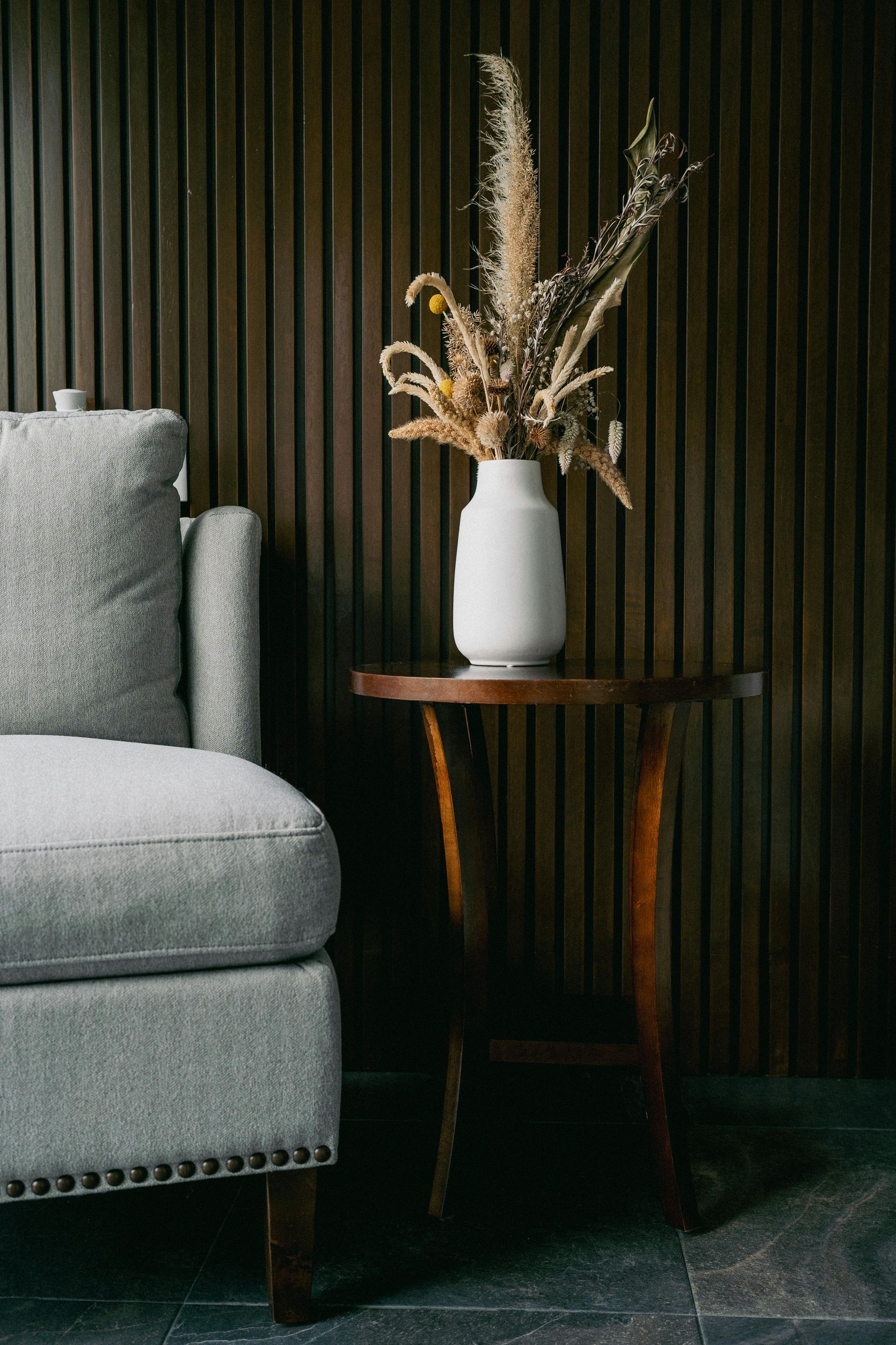 A gray armchair beside a dark wooden side table with a white vase holding dried flowers against a wood-paneled wall.
