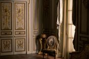Ornate room with gold detailing, light-colored walls, patterned chair, and large window with draped curtains.