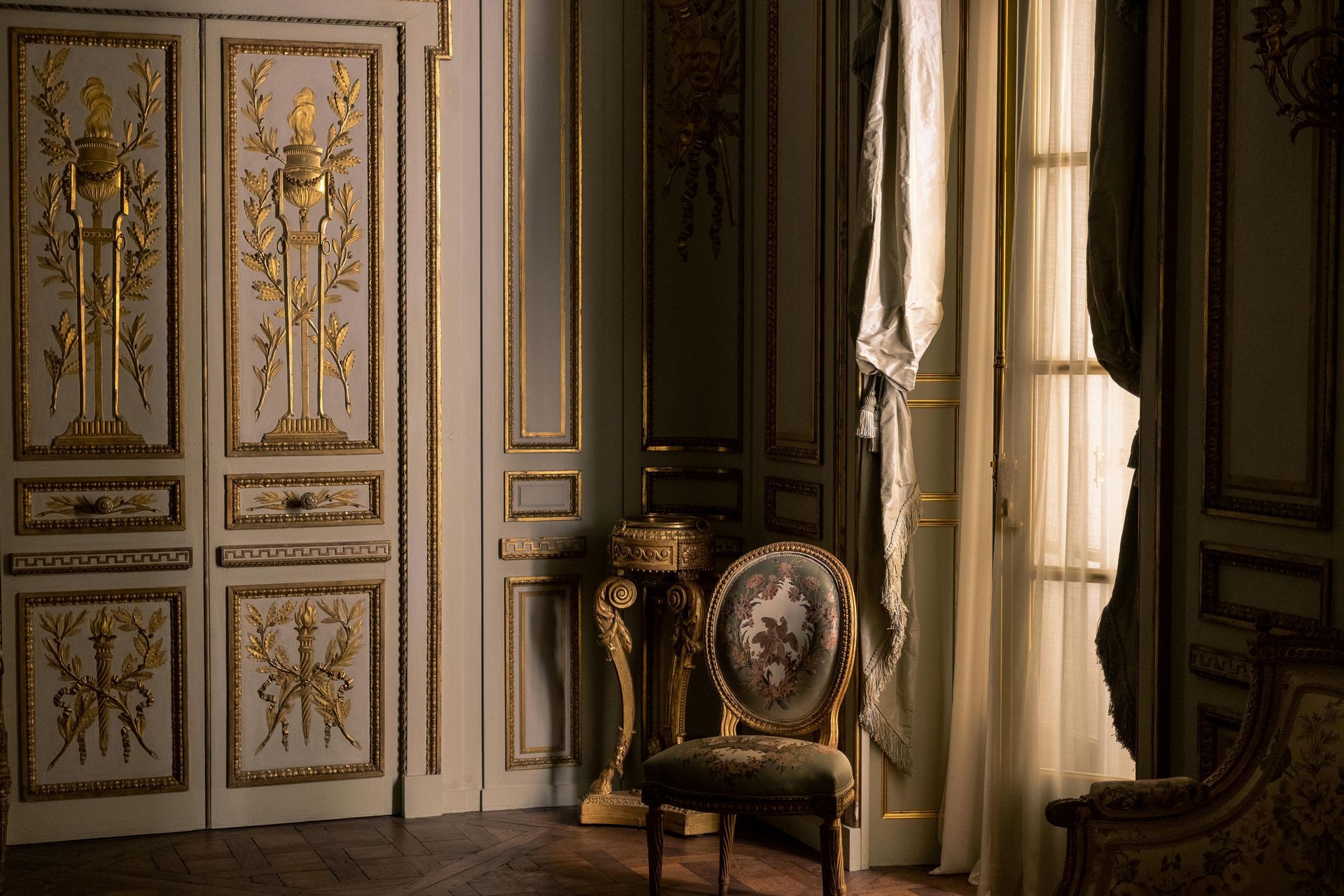 Ornate room with gold detailing, light-colored walls, patterned chair, and large window with draped curtains.