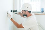 Construction worker in a white hard hat and gloves using a drill on a wall.