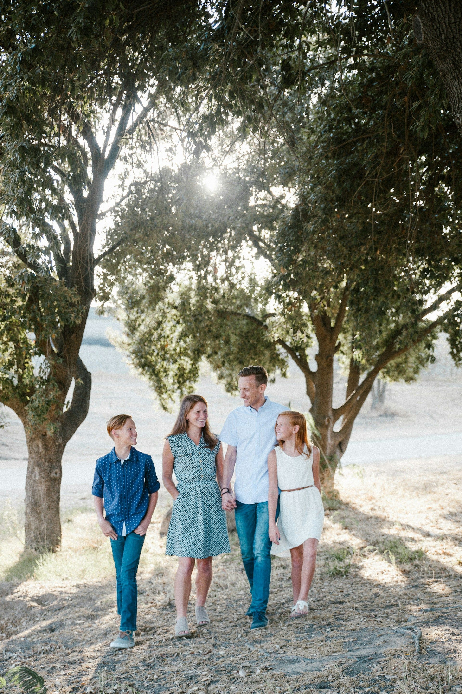 Four people walking under trees in a sunny park, with a dirt path and open field behind them.