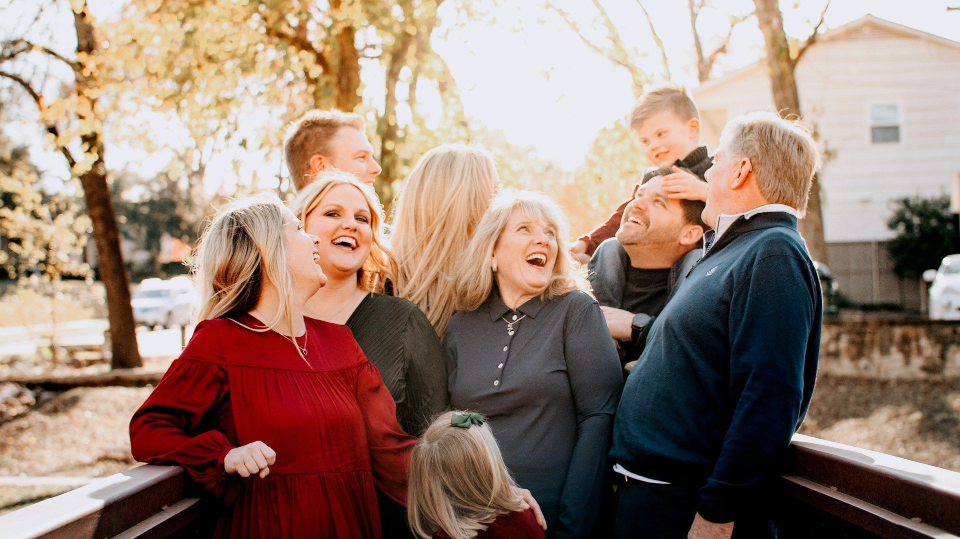 Family laughing together outdoors in warm sunlight, gathered by a wooden fence on a porch