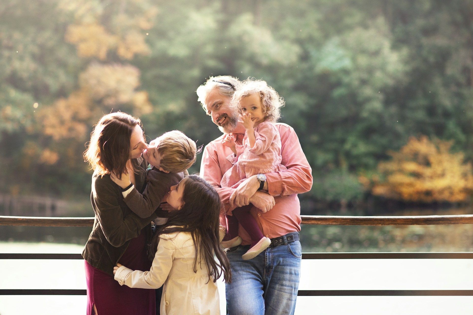 Four people hugging by a lakeside railing, with trees reflected in the water behind them.