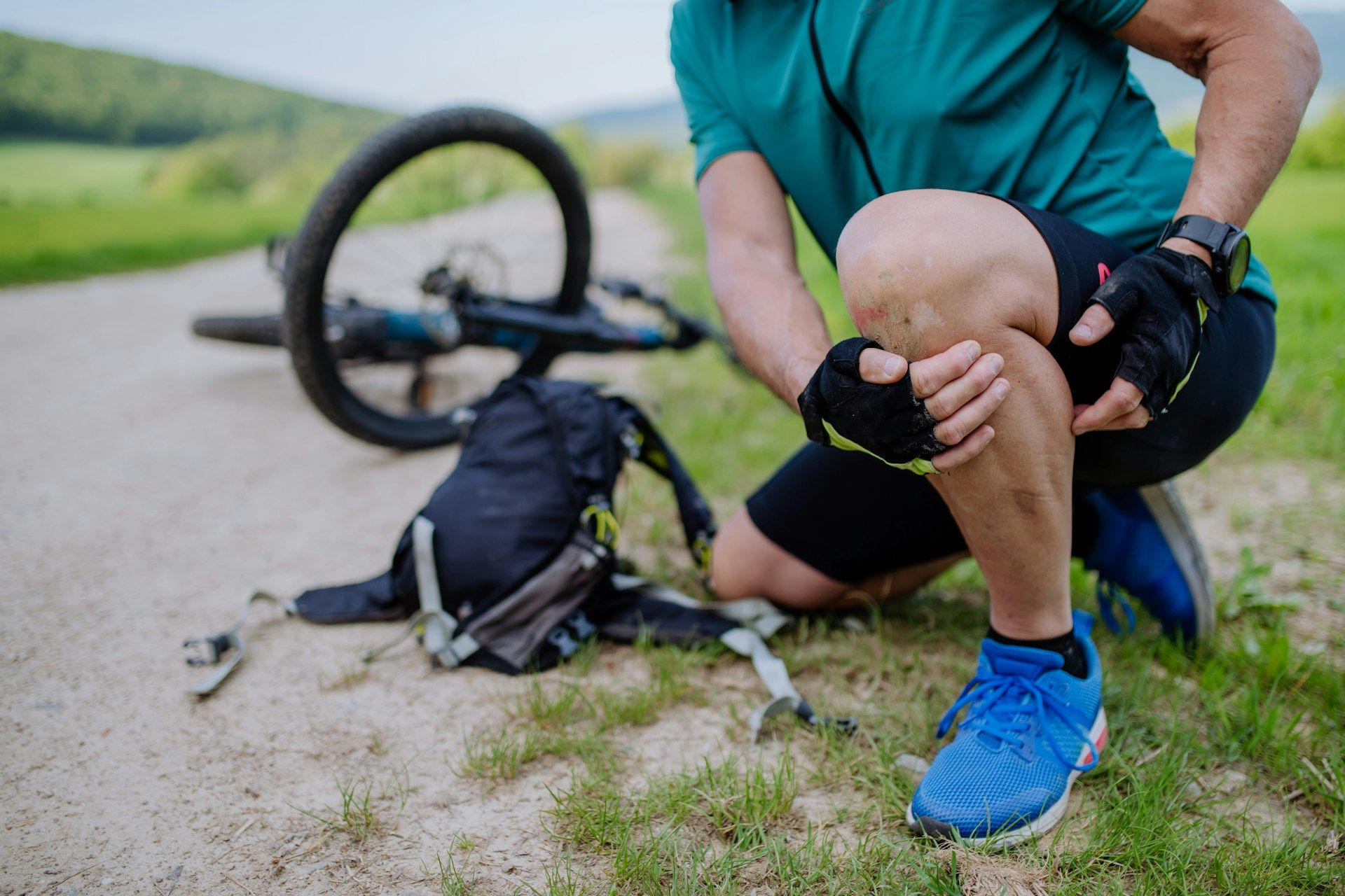 Cyclist kneeling beside a fallen bike on a gravel path, holding a sore knee with blue shoes visible