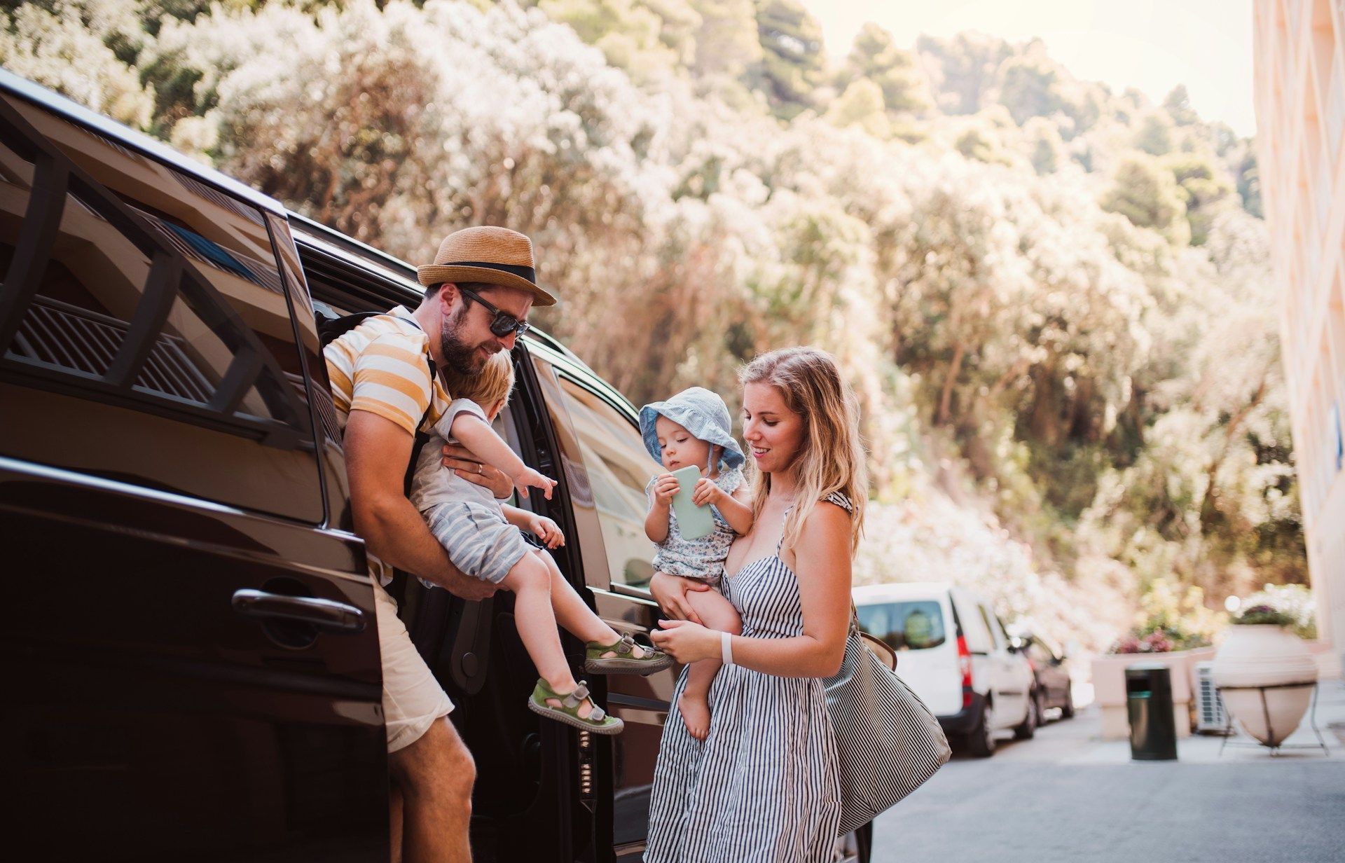 Family unloading bags from a van on a sunny roadside with parked cars and trees nearby