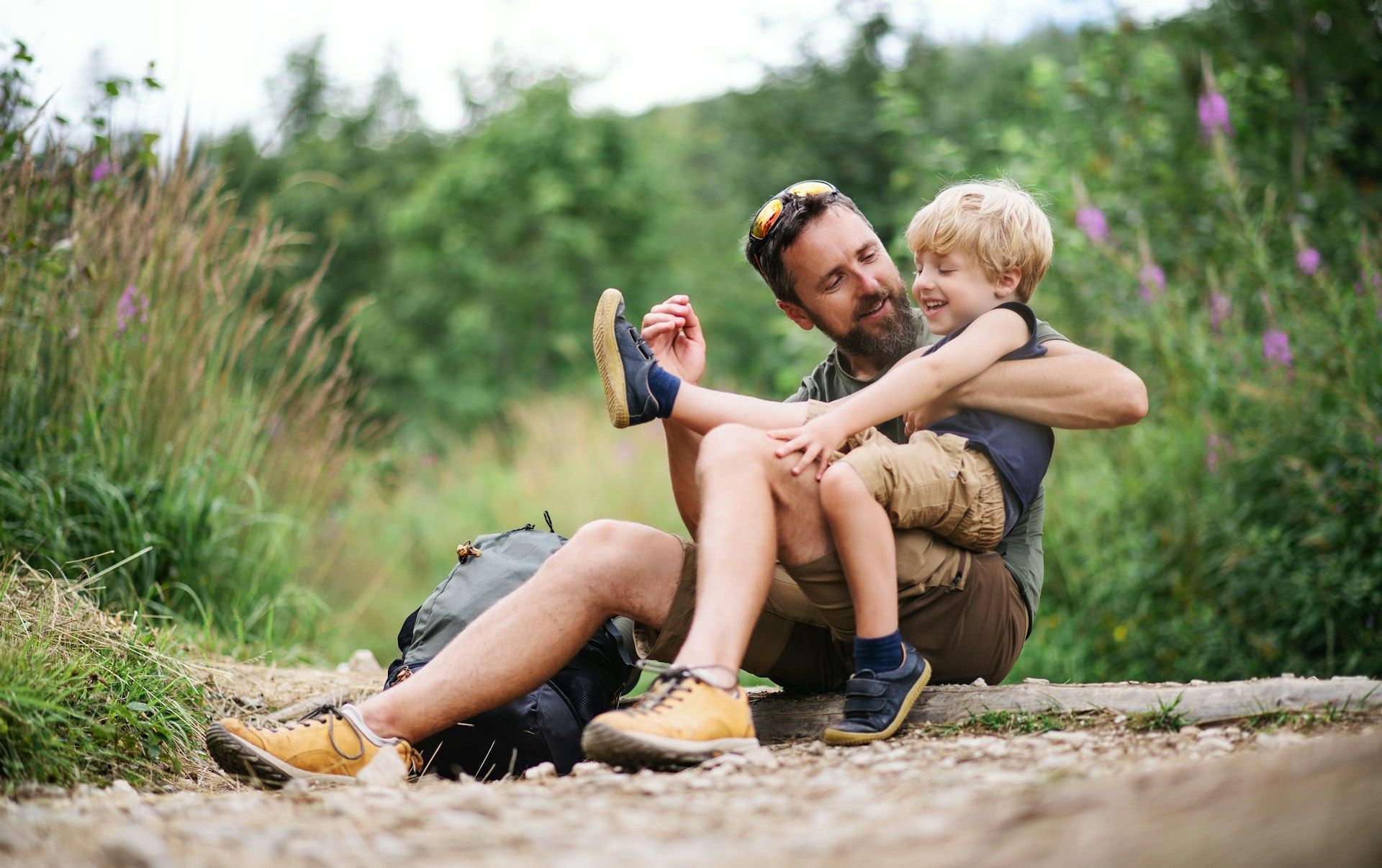 Man sitting on a path hugging a smiling child outdoors, both in casual clothes and boots.