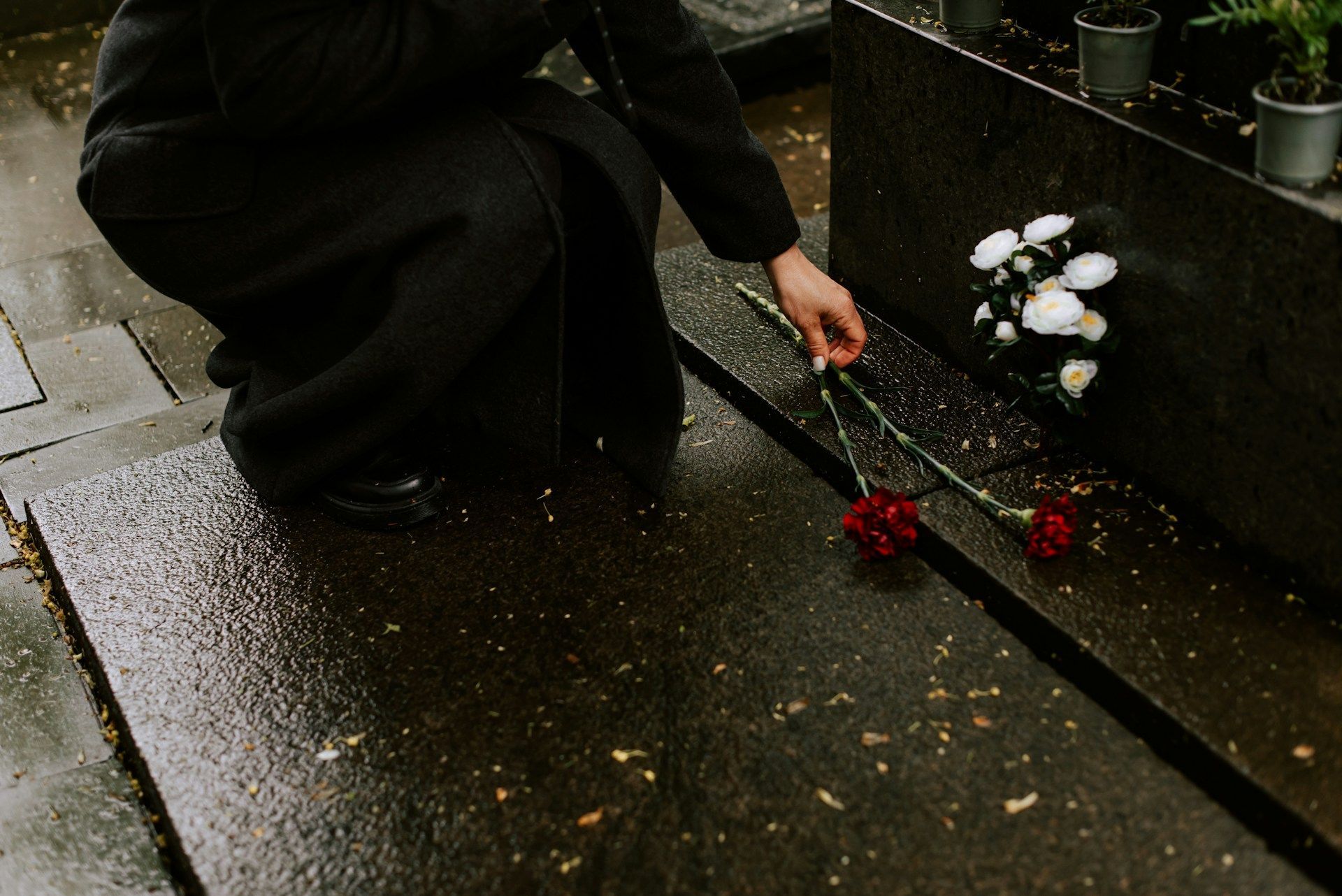 Person kneels by a wet graveside, placing red roses beside white flowers and candles.