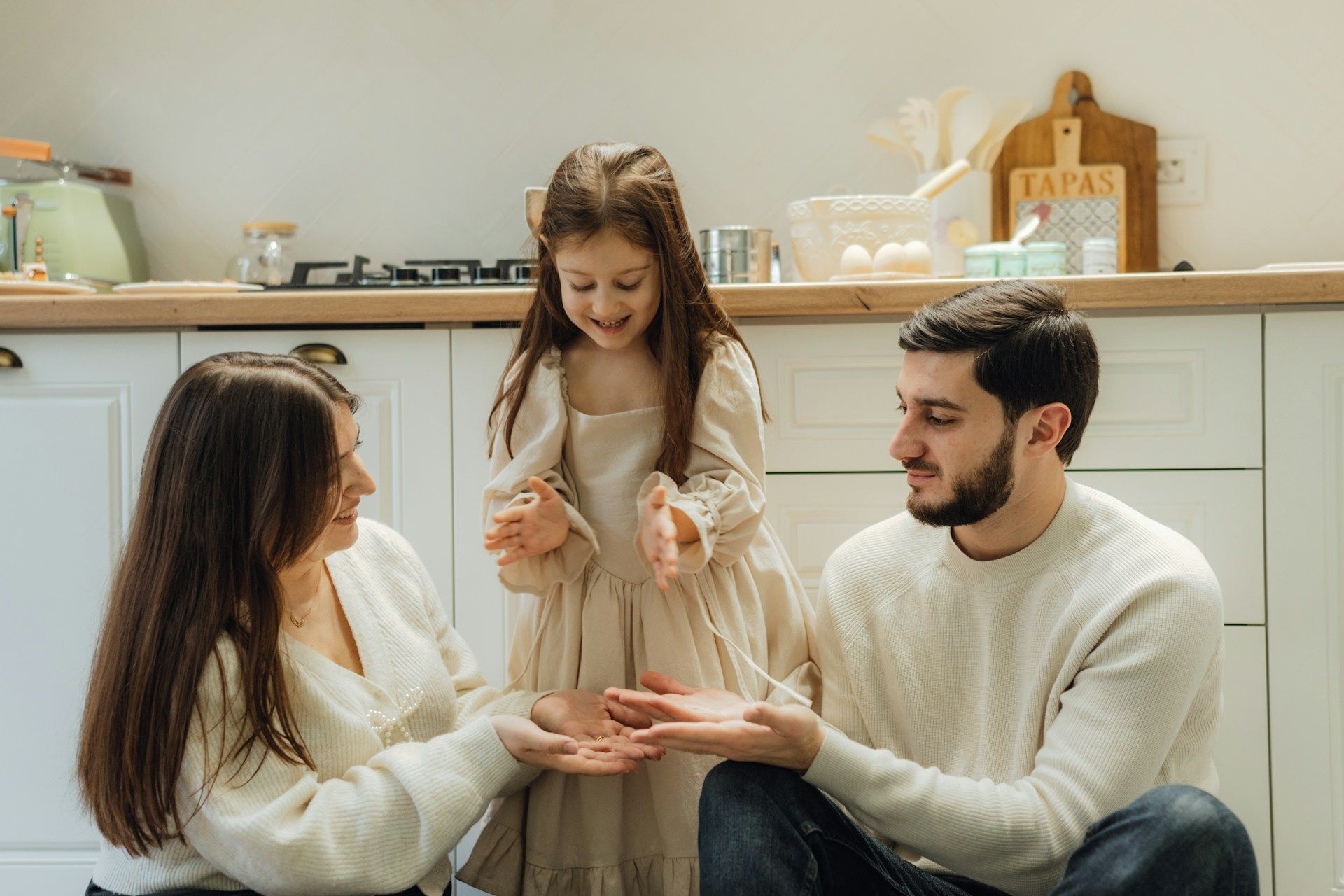 Family in a bright kitchen, smiling while a child stands between seated adults, holding their hands.