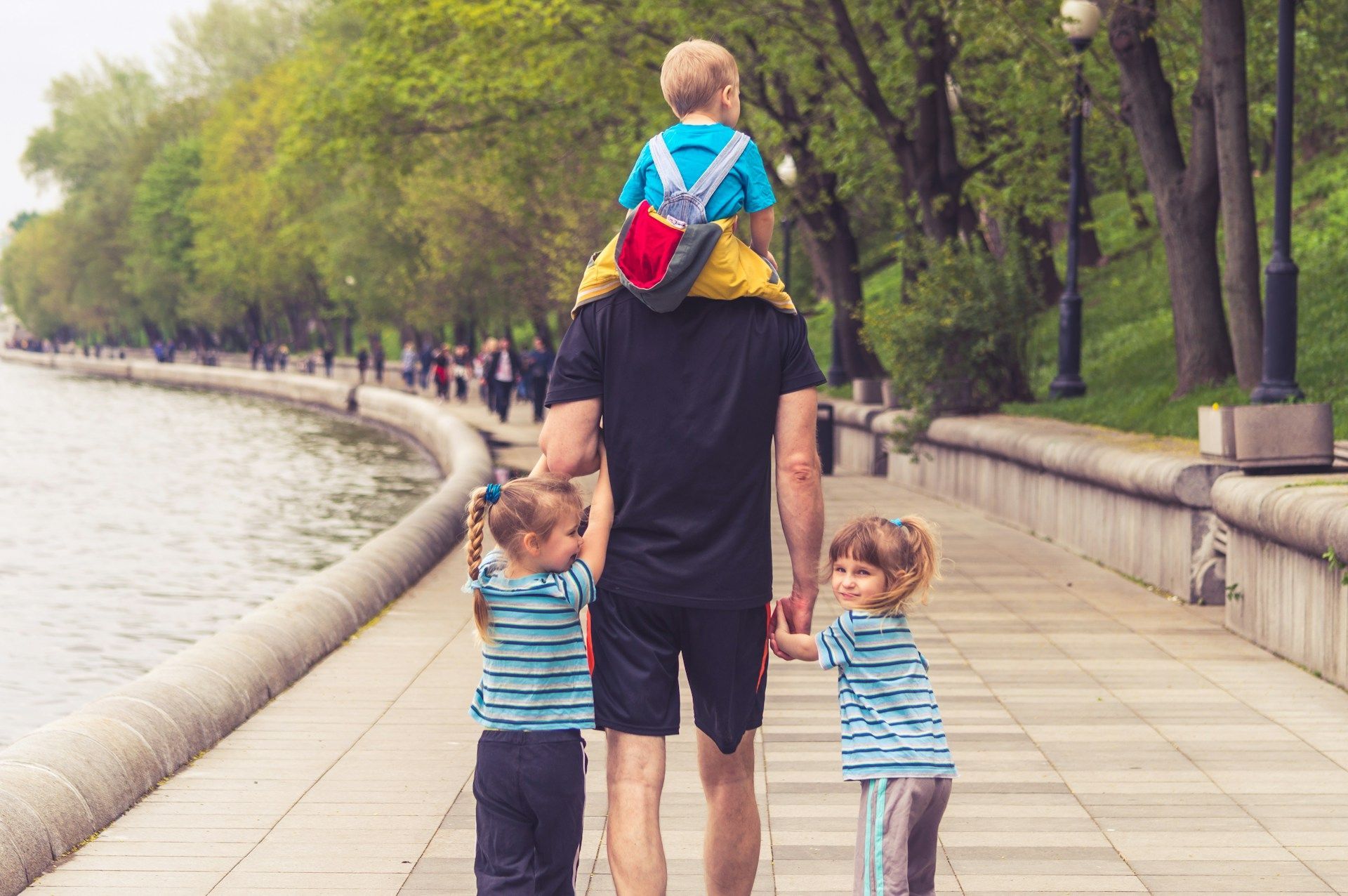 Person walking with two children along a riverside path, one child riding on their shoulders