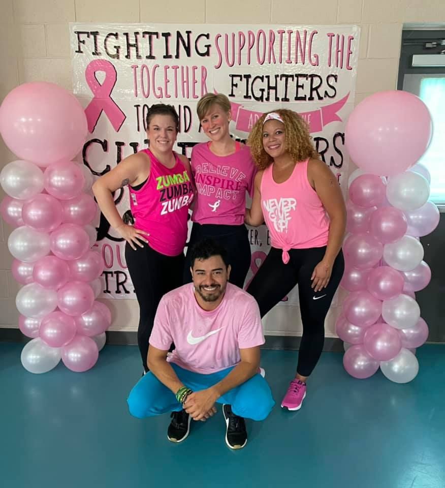 Group of people in pink, supporting breast cancer awareness. They are posing in front of pink balloons and a banner.