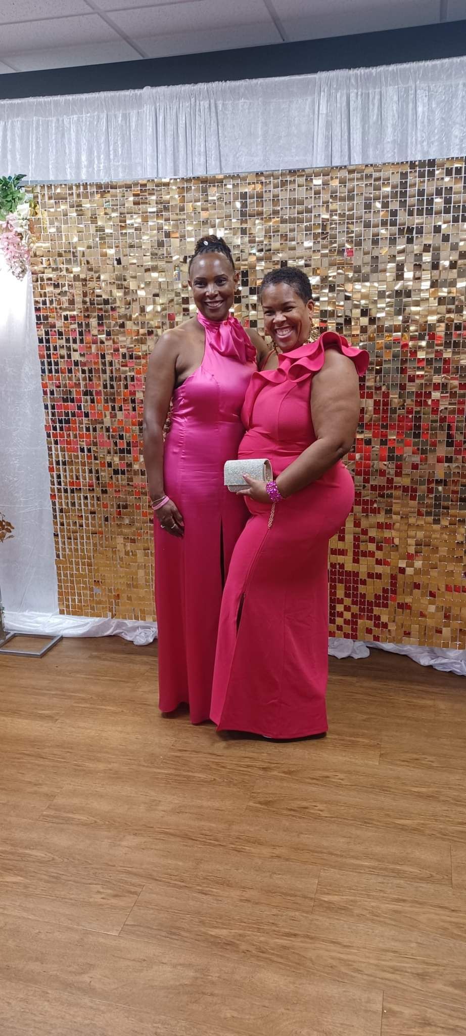 Two women in pink dresses stand smiling in front of a gold sequin backdrop.