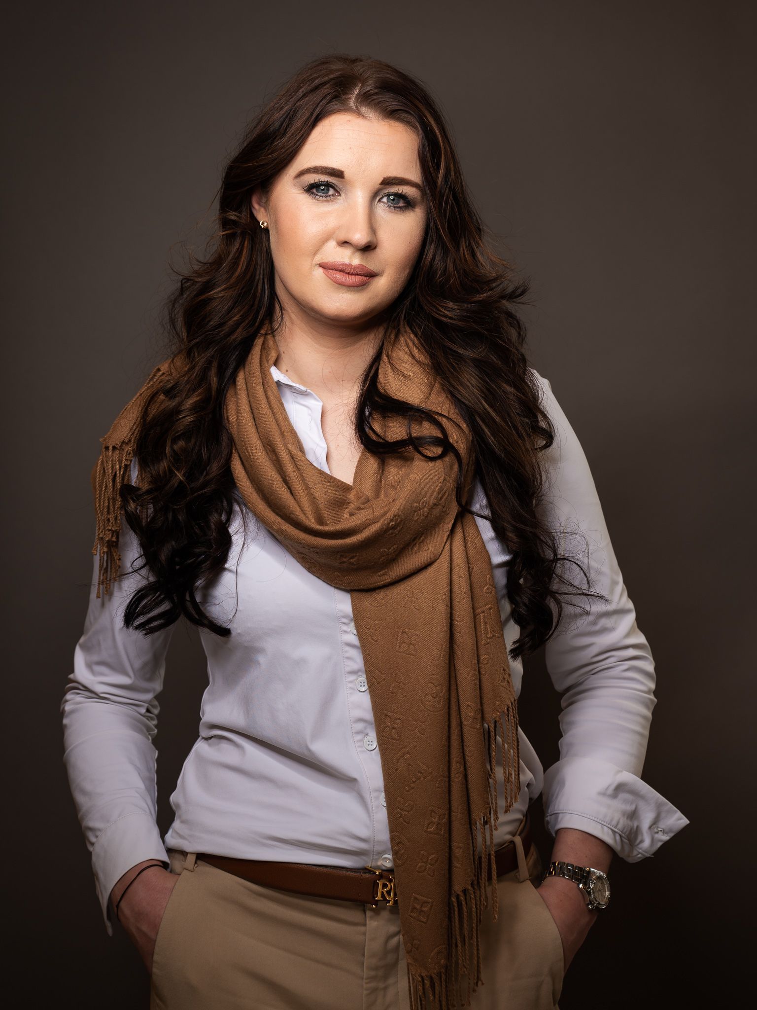Een vrouw draagt een wit shirt en een bruine sjaal en staat met haar handen in haar zakken.