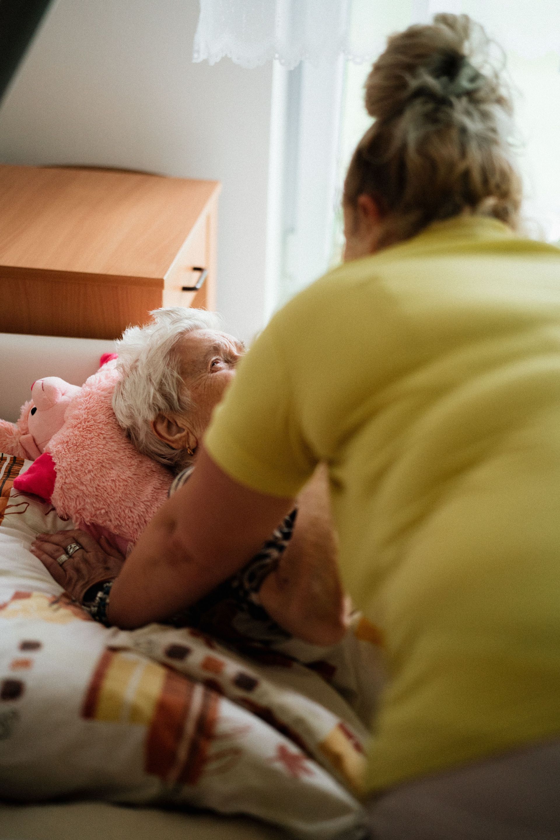 Person helping a small white dog on a bed in a softly lit room