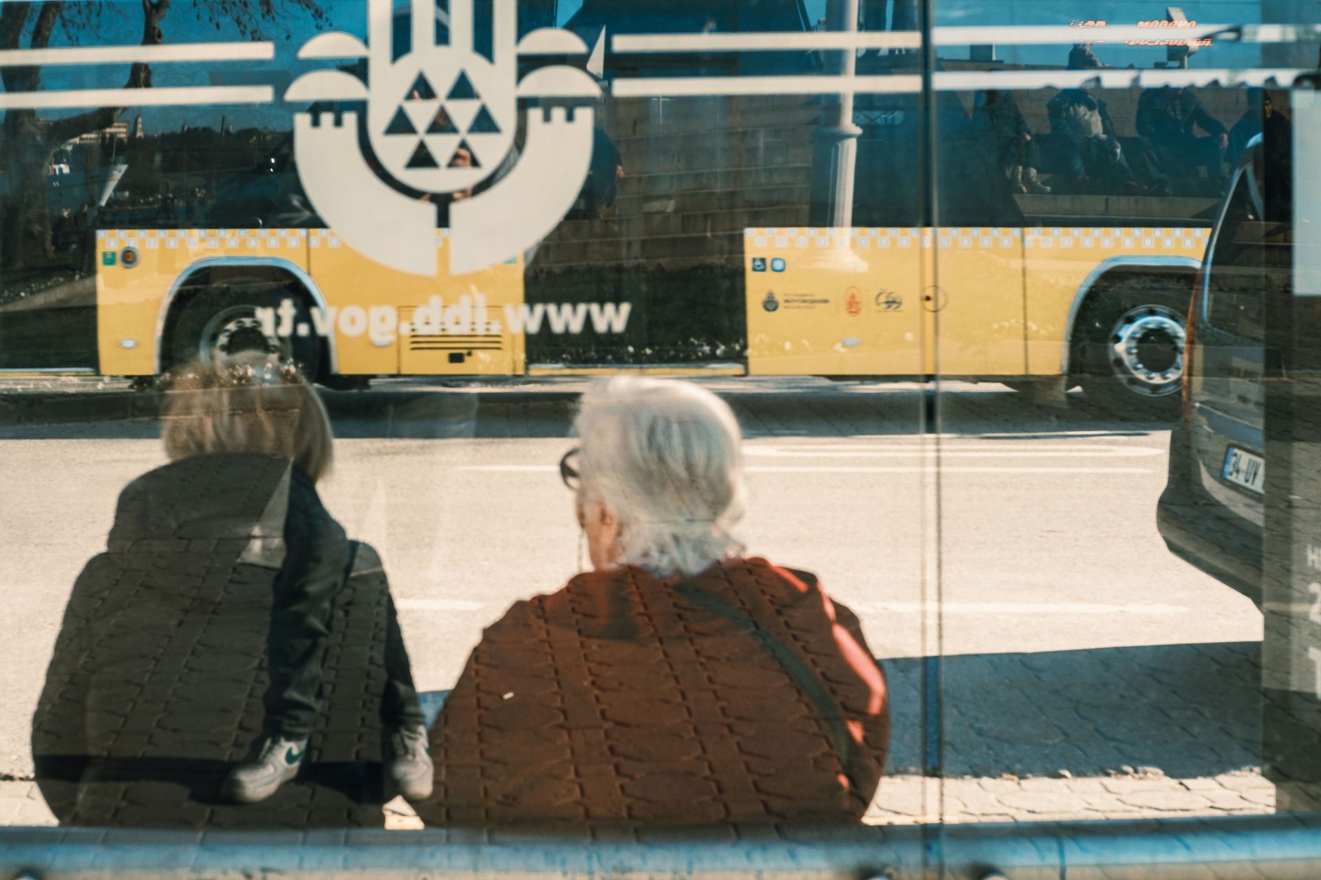 Two people sit at a bus stop, seen through glass, with a yellow bus passing behind them.